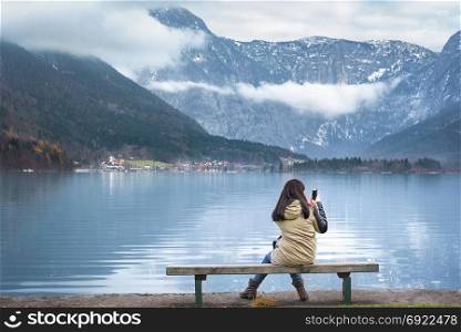 Woman sitting on a wooden bench, on the shores of the Hallstatter lake, using smartphone to photograph the Austrian Dachstein mountains, in Hallstatt, Austria.