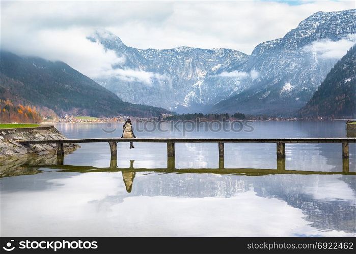 Woman sitting on a wooden bridge over the lake Hallstatter, admiring the peaks of the Austrian Alps and their reflection in the water, in Hallstatt, Austria.