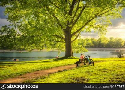 Woman sitting on bench and mountain bike, green trees and grass, lake at sunset in spring. Colorful landscape with resting girl, bicycle, river in park in summer. Sport and travel. Biking. Nature