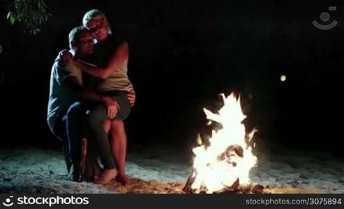 Woman sitting on man&acute;s knees and gently kissing her boyfriend. Couple enjoying romantic evening on the beach near bonfire and looking at fire