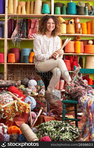 Woman Sitting On Stool Holding Knitting Needles In Front Of Yarn Display