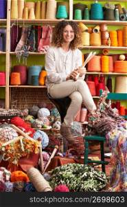 Woman Sitting On Stool Holding Knitting Needles In Front Of Yarn Display