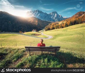 Woman sitting on the bench and beautiful alpine village at sunset in autumn. Dolomites, Italy. Colorful landscape with girl, green meadows, orange trees, road, mountain, sky, golden sunlight in fall