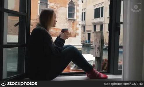 Woman sitting on the windowsill and having hot coffee. Venice canal and old worn buildings outside, boat sailing by