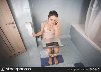 Woman sitting on toilet and using laptop and talking by phone