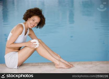 Woman sitting poolside using shower puff on leg smiling