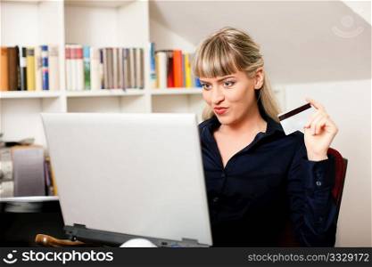 Woman sitting with a laptop in her home living room in front of a book shelf shopping or doing banking transactions online in the Internet, emphasized by shopping bags in the background and her holding a credit card