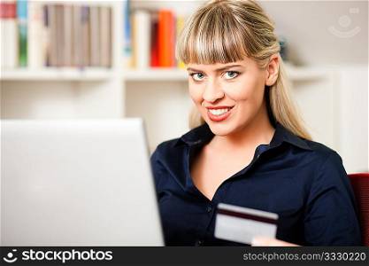 Woman sitting with a laptop in her home living room in front of a book shelf shopping or doing banking transactions online in the Internet, emphasized by shopping bags in the background and her holding a credit card