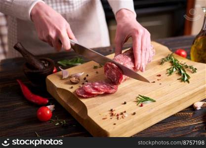 Woman slicing Traditional salami sausage on wooden cutting board by a knife at domestic kitchen.. Woman slicing Traditional salami sausage on wooden cutting board by a knife at domestic kitchen