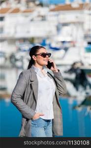 Woman smiles talking on her phone while taking a stroll at a coastal town on an out of focus background. Leisure concept.. Smiling woman talks on phone while strolling
