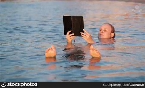 Woman smiling as she reads a book while swimming floating on her back in the water holding the book in the air