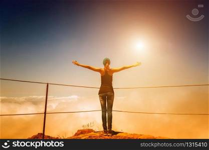 Woman standing back on cloudy mountain peak with fence at sunset, Portugal, Madeira. Woman on the mountain peak