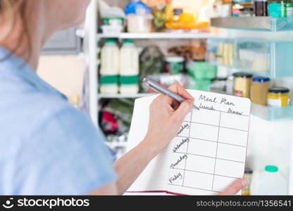 Woman Standing In Front Of Refrigerator In Kitchen With Notebook Writing Weekly Meal Plan