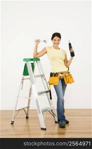 Woman standing in home with ladder and holding tools smiling.