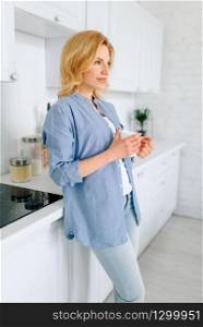 Woman standing on the kitchen with snow-white interior. Female person at home in the morning, healthy nutrition and lifestyle. Woman standing on kitchen with snow-white interior