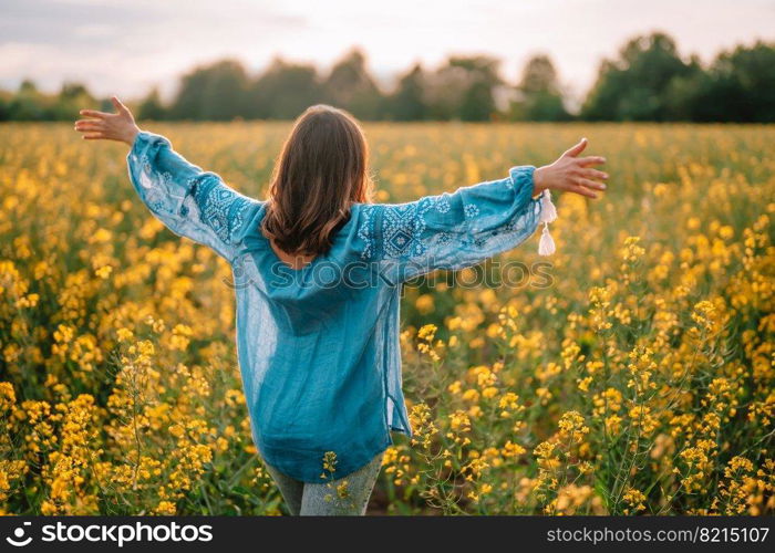 Woman standing with open arms in yellow canola field. Happy free lady in blue embroidered blouse. Alternative energy sources, oil production, biodiesel manufacturing, honey plant. High quality photo. Woman standing with open arms in yellow canola field. Happy free lady in blue embroidered blouse. Alternative energy sources, oil production, biodiesel manufacturing, honey plant.