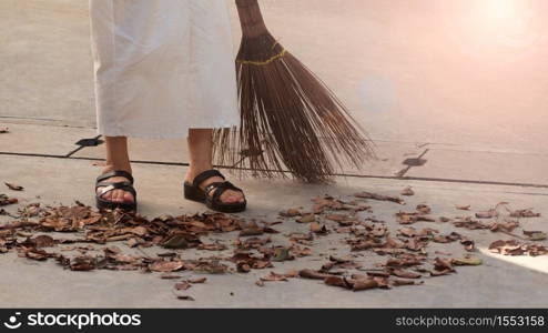 Woman sweeping dry leaves on the cement floor with long wood broom and keeping outdoor clean everyday which images showed middle aged female leg with white pants and black color sandal.