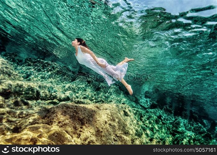Woman swimming near rock in transparent blue sea, wearing white dress, relaxation in refreshing water, summer enjoyment concept