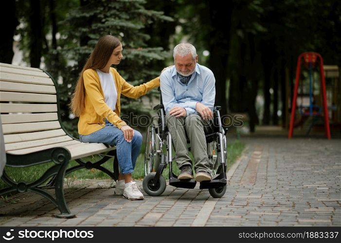 Woman takes care of a disabled father in wheelchair, family walking in park. Paralyzed people and disability, handicap overcoming. Handicapped male person and young female guardian in public place. Woman takes care of disabled father in wheelchair