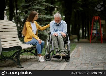Woman takes care of a disabled father in wheelchair, family walking in park. Paralyzed people and disability, handicap overcoming. Handicapped male person and young female guardian in public place. Woman takes care of disabled father in wheelchair
