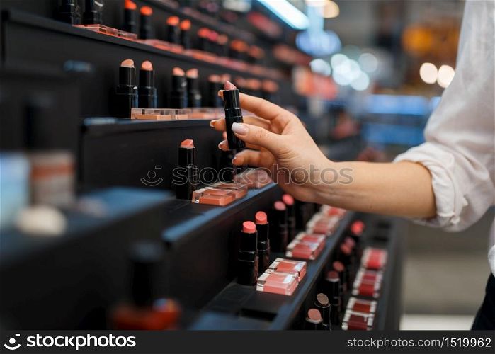 Woman takes lipstick from shelf in cosmetics store. Buyer at the showcase in luxury beauty shop salon, female customer in fashion market. Woman takes lipstick from shelf in cosmetics store