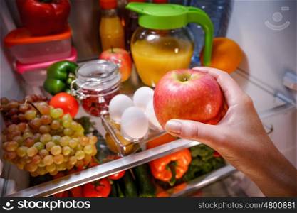 Woman takes the apple from the open refrigerator. Healthy food.