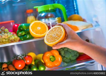 Woman takes the orange from the open refrigerator. Healthy food.