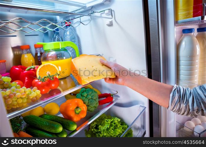 Woman takes the piece of cheese from the open refrigerator
