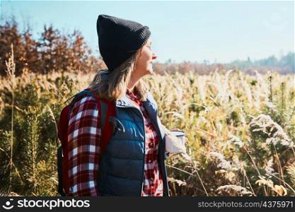 Woman taking break and enjoying the coffee during vacation trip. Woman standing on trail and looking at view. Woman with backpack hiking through tall grass along path in mountains. Spending summer vacation close to nature