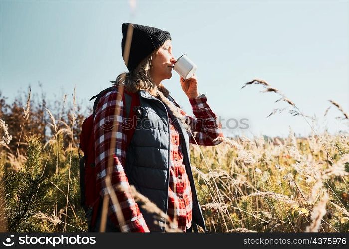 Woman taking break and enjoying the coffee during vacation trip. Woman standing on trail and looking away holding cup of coffee. Woman with backpack hiking through tall grass along path in mountains. Spending summer vacation close to nature