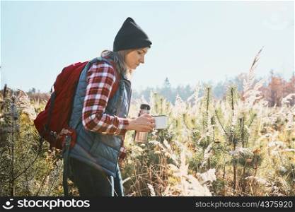 Woman taking break and relaxing in bright warm sunlight with cup of coffee during summer trip. Woman standing on trail and looking away. Woman with backpack hiking through tall grass along path in mountains. Spending summer vacation close to nature