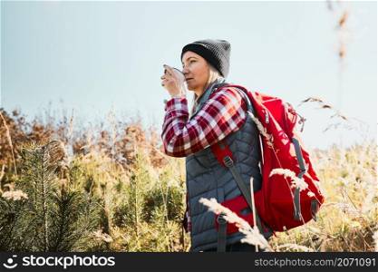 Woman taking break and relaxing with cup of coffee during summer trip. Woman standing on trail and looking away. Female with backpack hiking through tall grass along path in mountains. Spending summer vacation close to nature