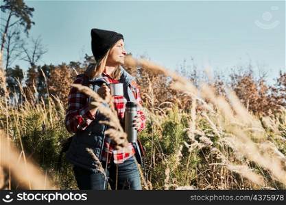 Woman taking break and relaxing with cup of coffee during summer trip. Woman standing on trail and looking away. Female with backpack hiking through tall grass along path in mountains. Spending summer vacation close to nature