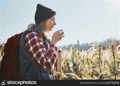 Woman taking break and relaxing with cup of coffee during summer trip. Woman standing on trail and looking away. Woman with backpack hiking through tall grass along path in mountains. Spending summer vacation close to nature
