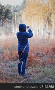 Woman taking photo of forest using camera in mobile phone. Forest in autumn season. Colorful foliage on trees lit by morning sunlight. Natural nature forest landscape in autumn warm sunlight day