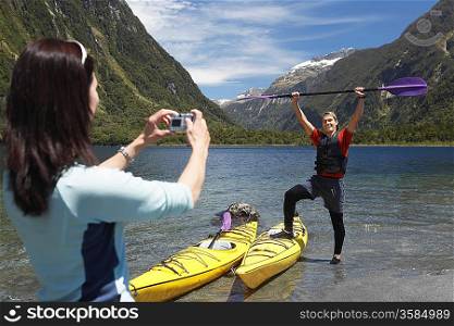 Woman taking picture of man hoisting oar of kayak over head on shore of mountain lake