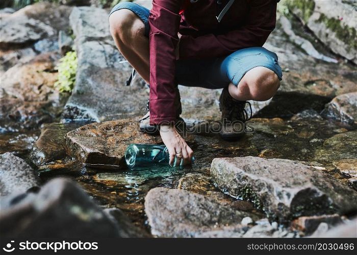 Woman taking pure water to bottle from mountain stream during trekking in mountains. Hiker crouching on rocks, filling bottle up with cold mountain water. Enjoying the outdoors in the summer trip vacation