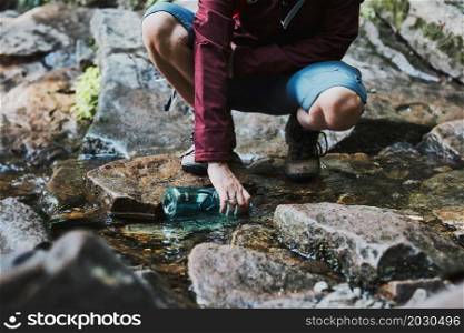 Woman taking pure water to bottle from mountain stream during trekking in mountains. Hiker crouching on rocks, filling bottle up with cold mountain water. Enjoying the outdoors in the summer trip vacation
