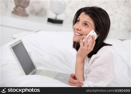 Woman talking on her mobile phone and using her laptop in bed