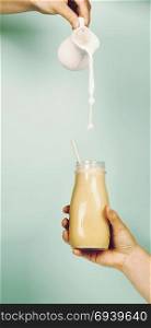 Woman (teenage girl) hand holding smoothie shake against bright wall. Pouring milk (cow or almond) into smoothie. Drinking healthy smoothie concept