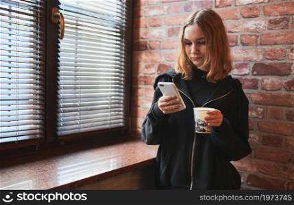 Woman texting on smartphone, drinking coffee while taking a break in office standing at window