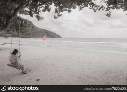 Woman. The phone is watching on a swing. Under a tree on the sea beach is crowded feel lonely.