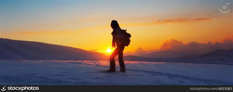 Woman traveler hiking in winter mountains, trekking in wintertime cold snowy weather, girl silhouette over natural colorful sky with bright sunset and beautiful landscape, freedom concept