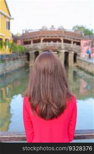 Woman traveler wearing Ao Dai Vietnamese dress sightseeing at Japanese covered bridge in Hoi An town, Vietnam. landmark and popular for tourist attractions. Vietnam and Southeast Asia travel concept