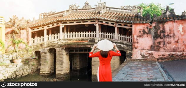 Woman traveler wearing Ao Dai Vietnamese dress sightseeing at Japanese covered bridge in Hoi An town, Vietnam. landmark and popular for tourist attractions. Vietnam and Southeast Asia travel concept