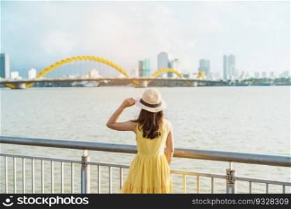 Woman Traveler with yellow dress visiting in Da Nang city. Tourist sightseeing the river view with Dragon bridge. Landmark and popular for tourist attraction. Vietnam and Southeast Asia travel concept