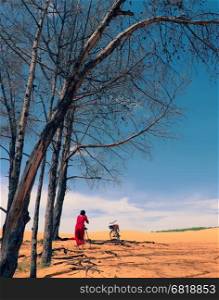 Woman traveling alone at Mui Ne, Vietnam, girl in red dress and selfie with bike on yellow sand hill under tree, tourist enjoy the trip in summetime