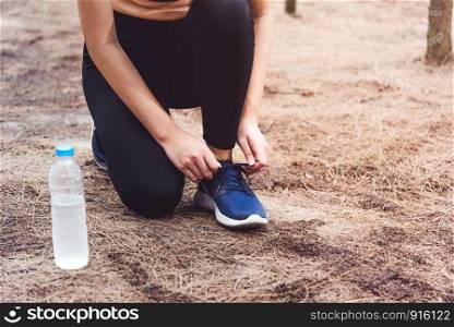 Woman tying up shoelaces when jogging in forest back with drinking water bottle beside hers. Sneakers rope tying. People and lifestyles concept. Healthcare and Wellness theme. Park and Outdoors theme.