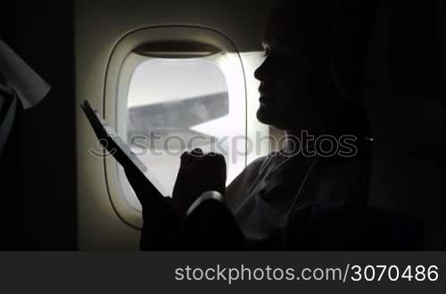 Woman typing on tablet computer sitting by illuminator in plane. Filling in time during the flight