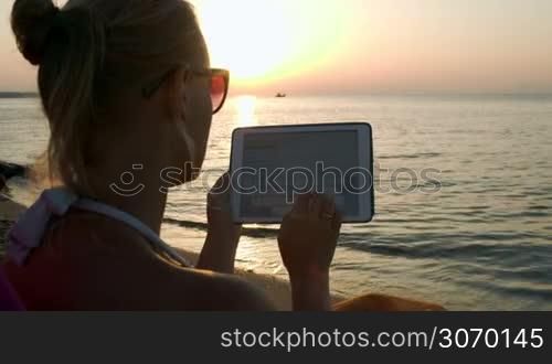 Woman typing on tablet computer sitting on beach at sunset. Vacation with pad